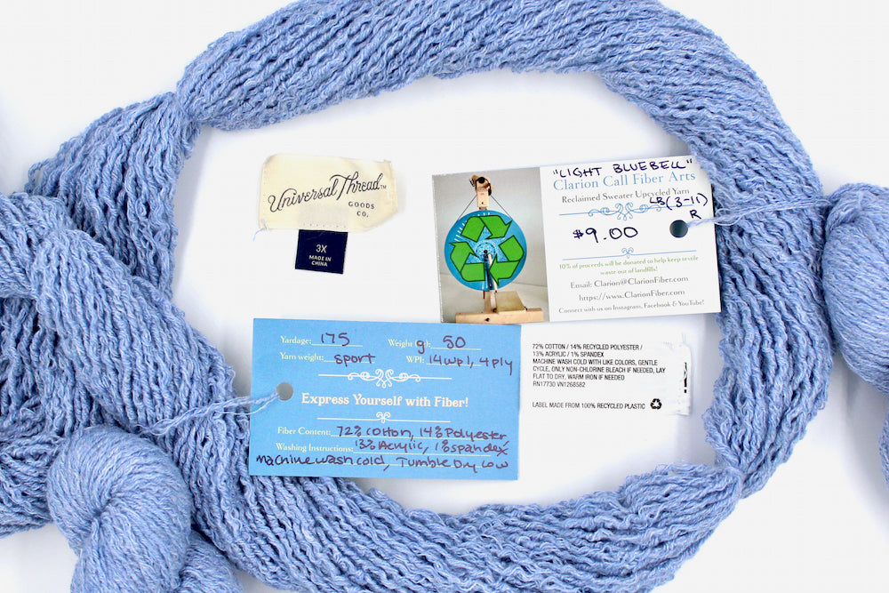 A skein of Vegan, baby blue, upcycled Cotton, Polyester, and acrylic, Sport Weight yarn encircling the sweater labels it was recycled from, as well as the hand written yarn tags detailing the specs of this recycled yarn to help you make your next project!
