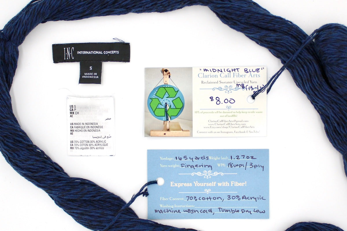 A skein of Vegan, Navy Blue/ Black, Cotton/ Acrylic, Fingering Weight yarn encircling the sweater labels it was recycled from, as well as the hand written yarn tags detailing the specs of this upcycled yarn to help you make your next project!