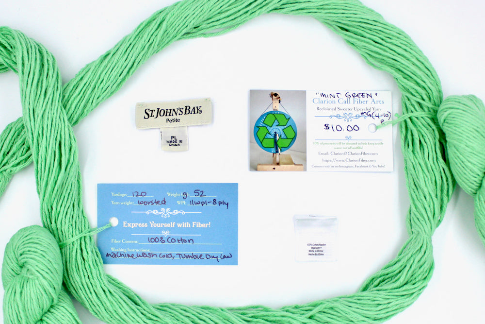 A skein of Vegan, vibrant light green, 100% Cotton, Worsted Weight yarn encircling the sweater labels it was recycled from, as well as the hand written yarn tags detailing the specs of this recycled yarn to help you make your next project!