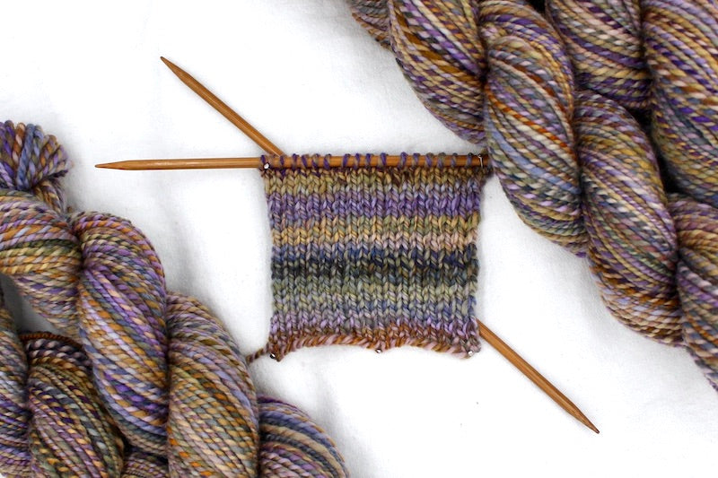A sample swatch knitted from a one of a kind, hand dyed variegated skein of multicolored Purple, Lavender, Periwinkle, Taupe, Gold, and Orange self-striping wool Yarn.