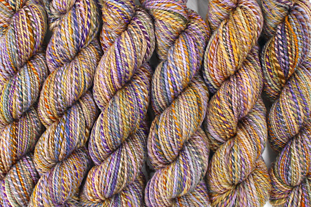 Six one of a kind, Dk Weight, hand dyed Variegated skeins of multicolored Purple, Lavender, Periwinkle, Taupe, Gold, and Orange self-striping Yarn, made of 100% Hand Dyed Merino Wool lined up side by side.