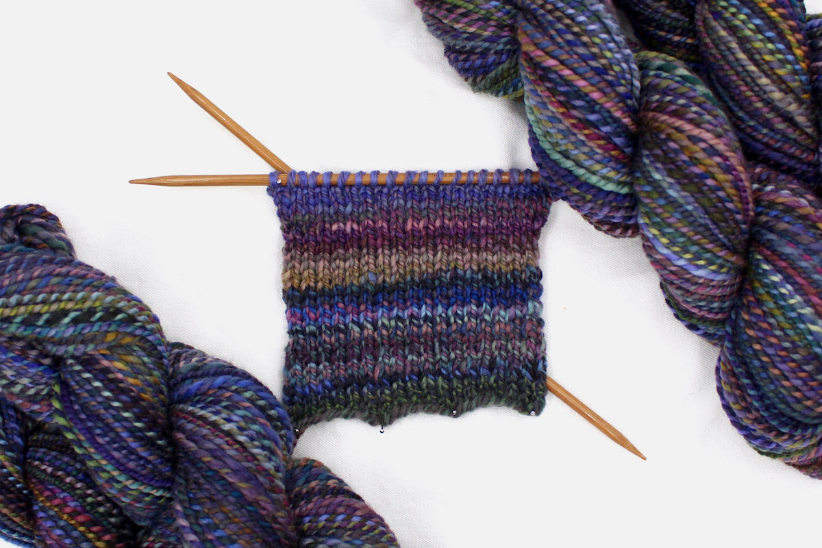 A sample swatch knitted from a one of a kind, Worsted weight, hand dyed variegated skein of multicolored Blue, Purple, Yellow, and Olive Green, and Maroon self-striping wool Yarn.