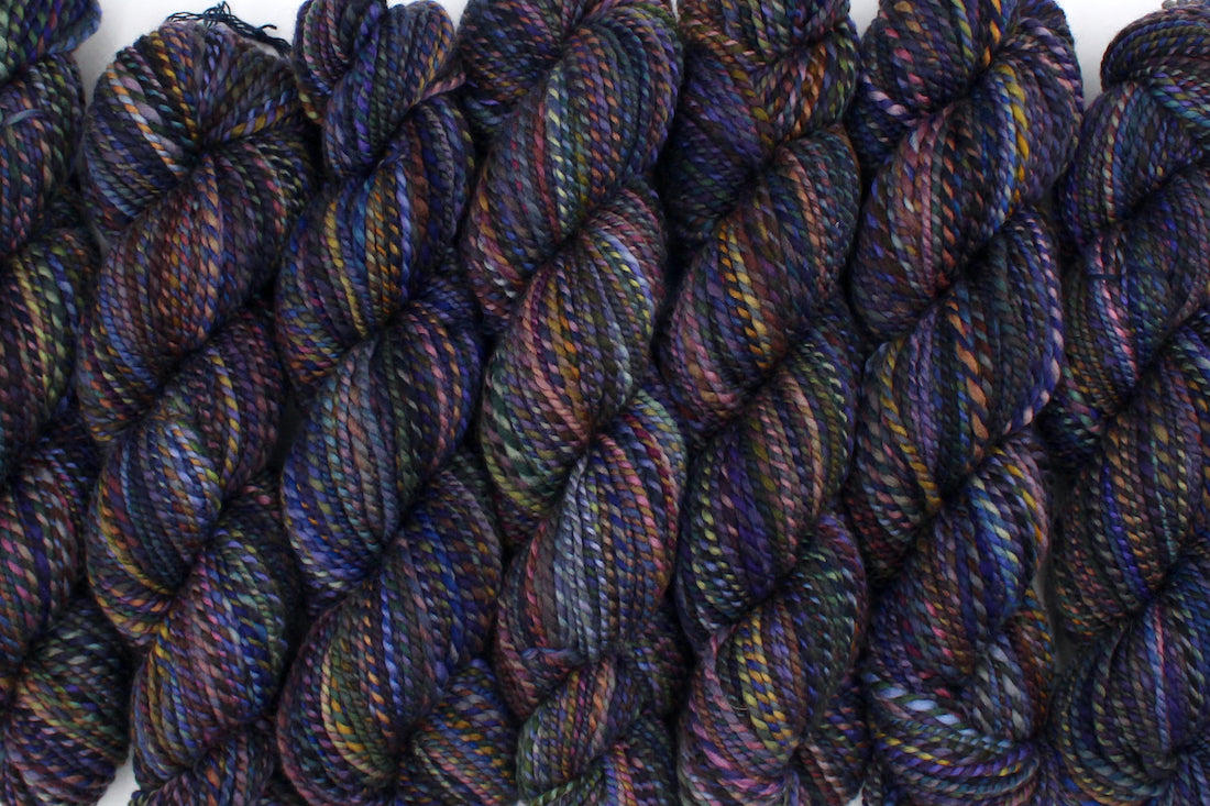 Six one of a kind, Worsted weight, hand dyed variegated skein of multicolored Blue, Purple, Yellow, and Olive Green, and Maroon self-striping wool Yarn lined up side by side.