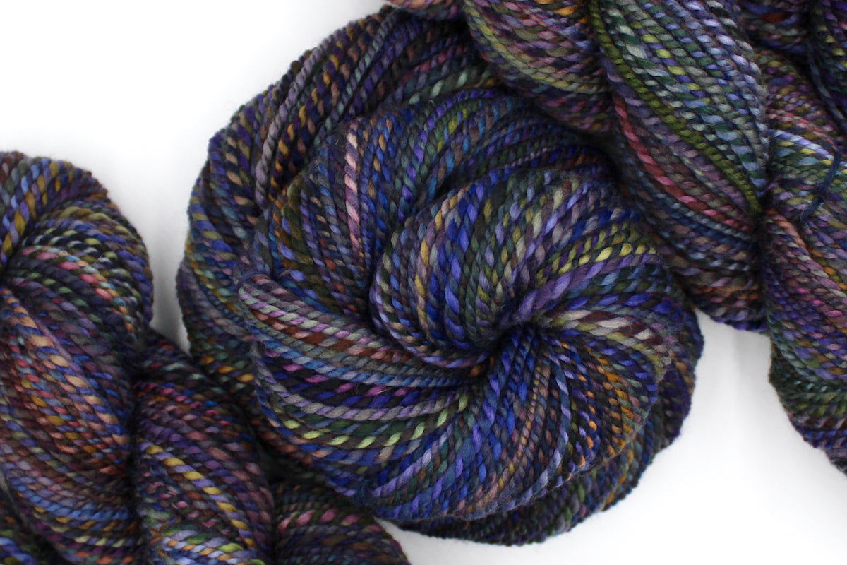 A one of a kind, Worsted Weight, hand dyed variegated skein of multicolored Blue, Purple, Yellow, and Olive Green, and Maroon, self-striping wool Yarn coiled attractively in the center of the frame.