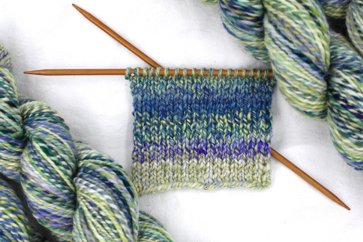A sample swatch knitted from a one of a kind, Worsted weight, hand dyed variegated skein of multicolored Blue, Green, Teal, and Light Yellow self-striping wool Yarn.