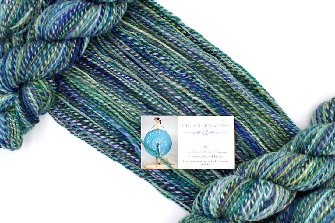 A one of a kind, Worsted weight, hand dyed variegated skein of multicolored Blue, Green, Teal, and Light Yellow self-striping wool Yarn draped diagonally across the frame, so you can really see the color play.