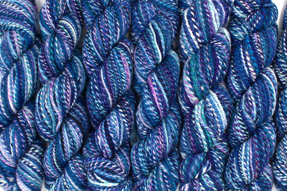 Six one of a kind, Dk Weight, hand dyed variegated skeins of multicolored Blue, and Purple self-striping Hand spun Yarn, made of 100% Hand Dyed Merino Wool lined up side by side.