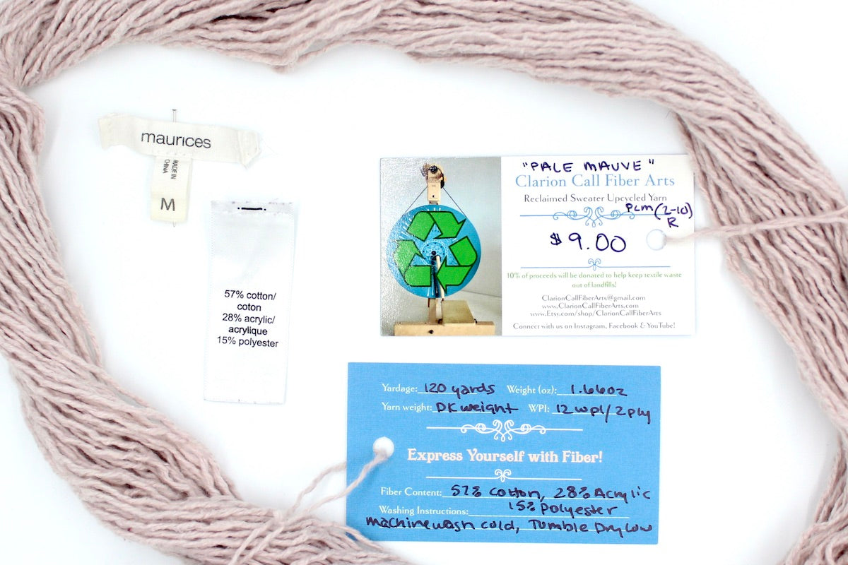 A skein of Vegan, Light Pastel Pink, Cotton/ Acrylic/ Polyester, Dk Weight yarn encircling the sweater labels it was recycled from, as well as the hand written yarn tags detailing the specs of this upcycled yarn to help you make your next project!