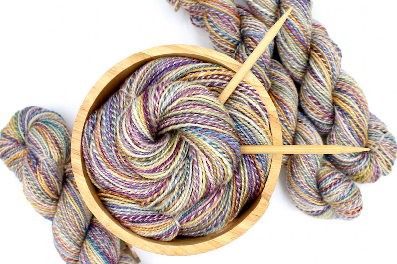 A one of a kind, hand dyed variegated skein of multicolored Purple, Pink, Blue, Yellow and Orange self-striping wool Yarn, in a yarn bowl with knitting needles, ready to be made into something beautiful!