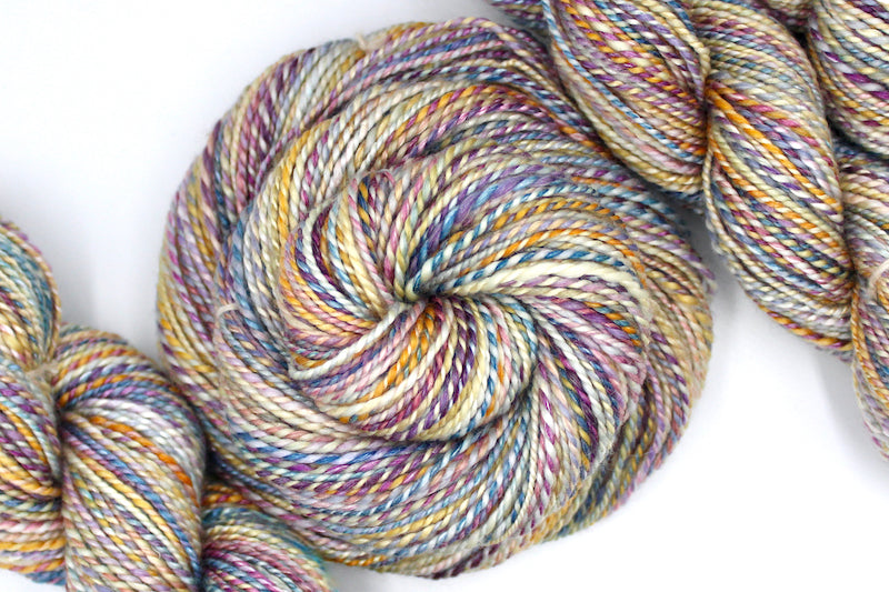 A one of a kind, hand dyed variegated skein of multicolored Purple, Pink, Blue, Yellow and Orange self-striping wool Yarn coiled attractively in the center of the frame.