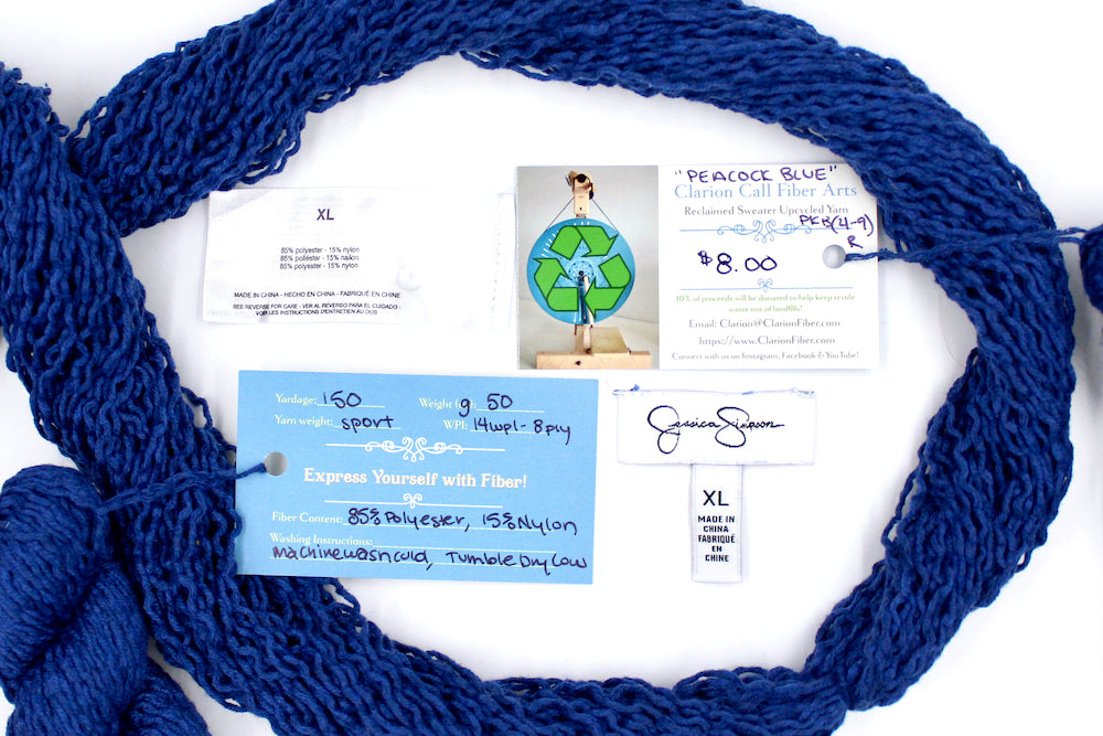 A skein of Vegan, Purplish Blue, 85% Polyester, 15% Nylon, Sport Weight yarn encircling the sweater labels it was recycled from, as well as the hand written yarn tags detailing the specs of this recycled yarn to help you make your next project!