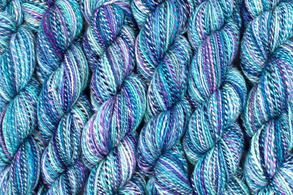Six one of a kind, Dk Weight, hand dyed variegated skeins of multicolored Blue, Pink, and Purple self-striping Hand spun Yarn, made of 100% Hand Dyed Superwash Merino Wool lined up side by side.
