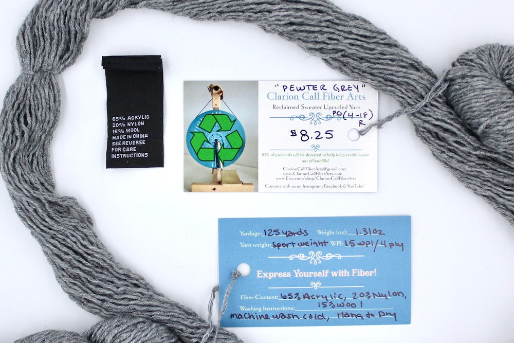 A skein of Silver Grey, Acrylic/ Nylon/ Wool, Sport Weight yarn encircling the sweater labels it was recycled from, as well as the hand written yarn tags detailing the specs of this upcycled yarn to help you make your next project!