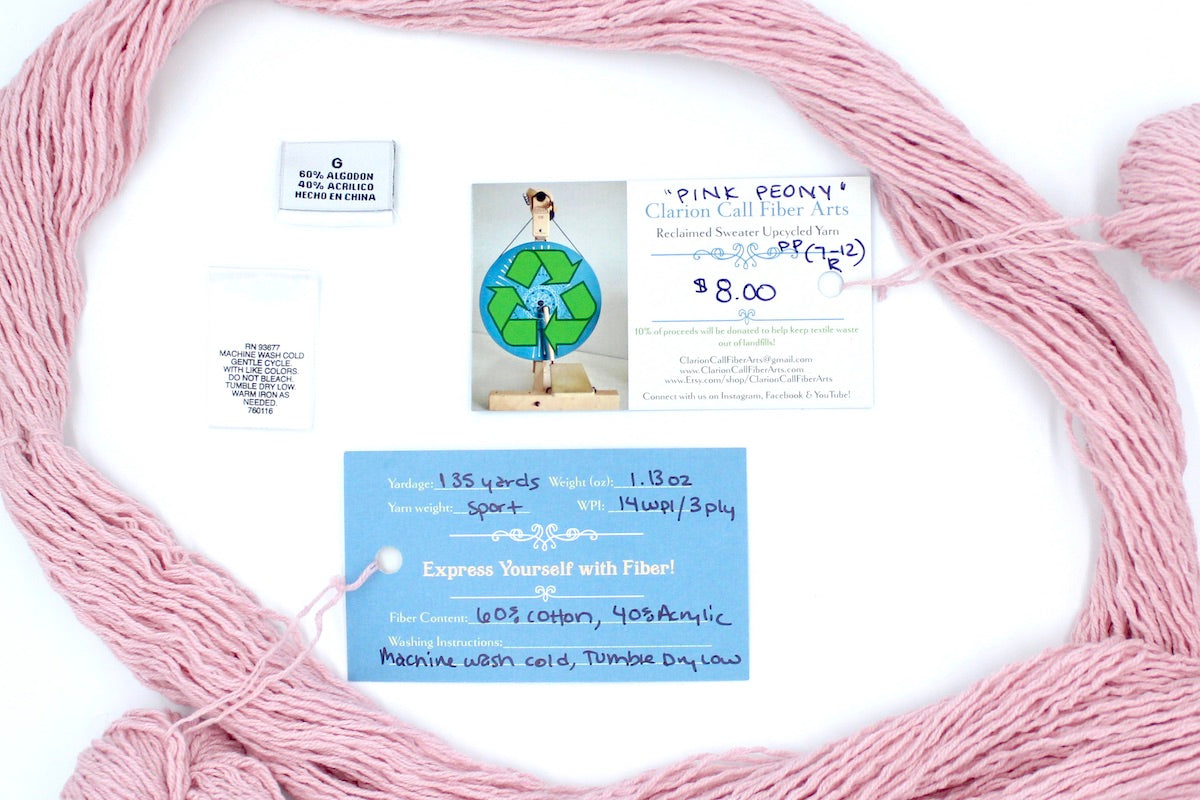 A skein of Vegan, Light Ballet Pink, Cotton/ Acrylic, Sport Weight yarn encircling the sweater labels it was recycled from, as well as the hand written yarn tags detailing the specs of this upcycled yarn to help you make your next project!