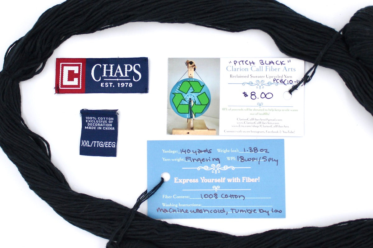 A skein of Vegan, Black, 100% Cotton, Fingering Weight yarn encircling the sweater labels it was recycled from, as well as the hand written yarn tags detailing the specs of this upcycled yarn to help you make your next project!