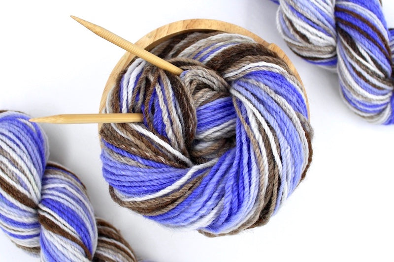 A one of a kind, hand dyed Variegated skein of multicolored Purple, Lavender, White, Tan, and Brown self-striping wool Yarn, in a yarn bowl with knitting needles, ready to be made into something beautiful!
