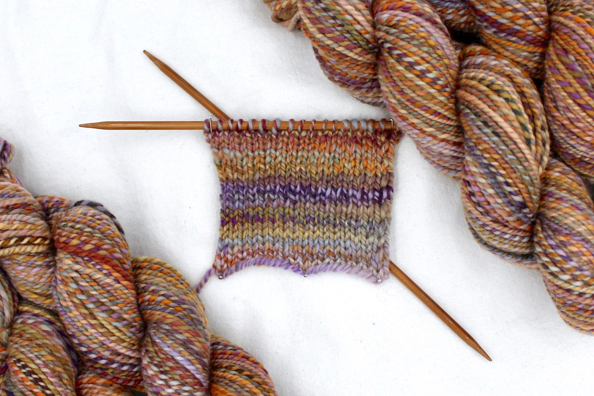 A sample swatch knitted from a one of a kind, Worsted weight, hand dyed variegated skein of multicolored Orange, Yellow, Pink, Purple, Olive Green, and Blue self-striping wool Yarn.