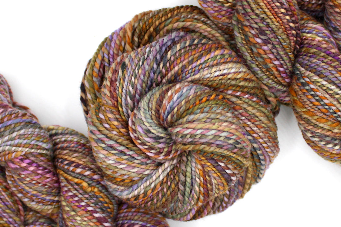 A one of a kind, Worsted Weight, hand dyed variegated skein of multicolored Orange, Yellow, Pink, Purple, Olive Green, and Blue, self-striping wool Yarn coiled attractively in the center of the frame.
