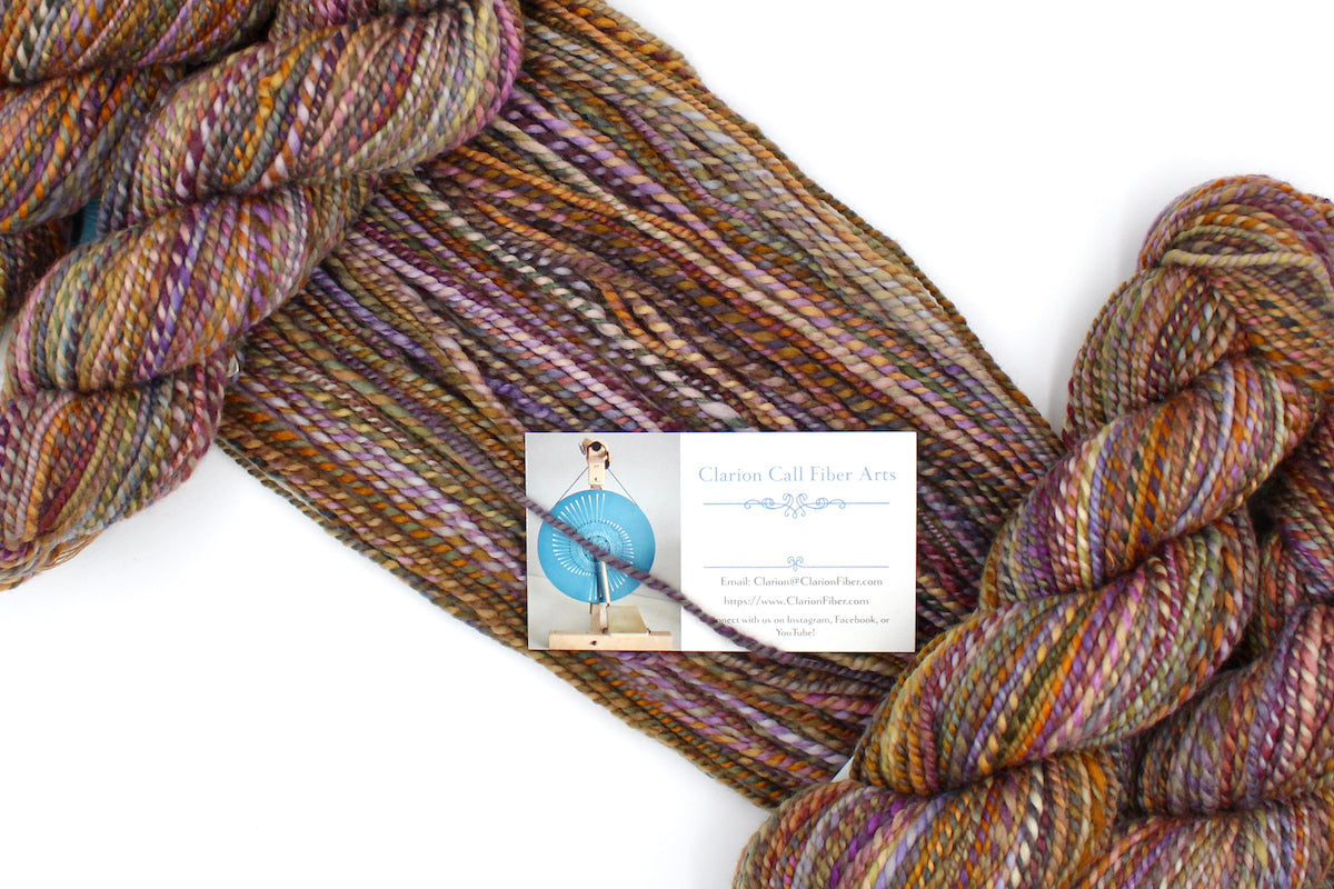 A one of a kind, Worsted weight, hand dyed variegated skein of multicolored Orange, Yellow, Pink, Purple, Olive Green, and Blue self-striping wool Yarn draped diagonally across the frame, so you can really see the color play.