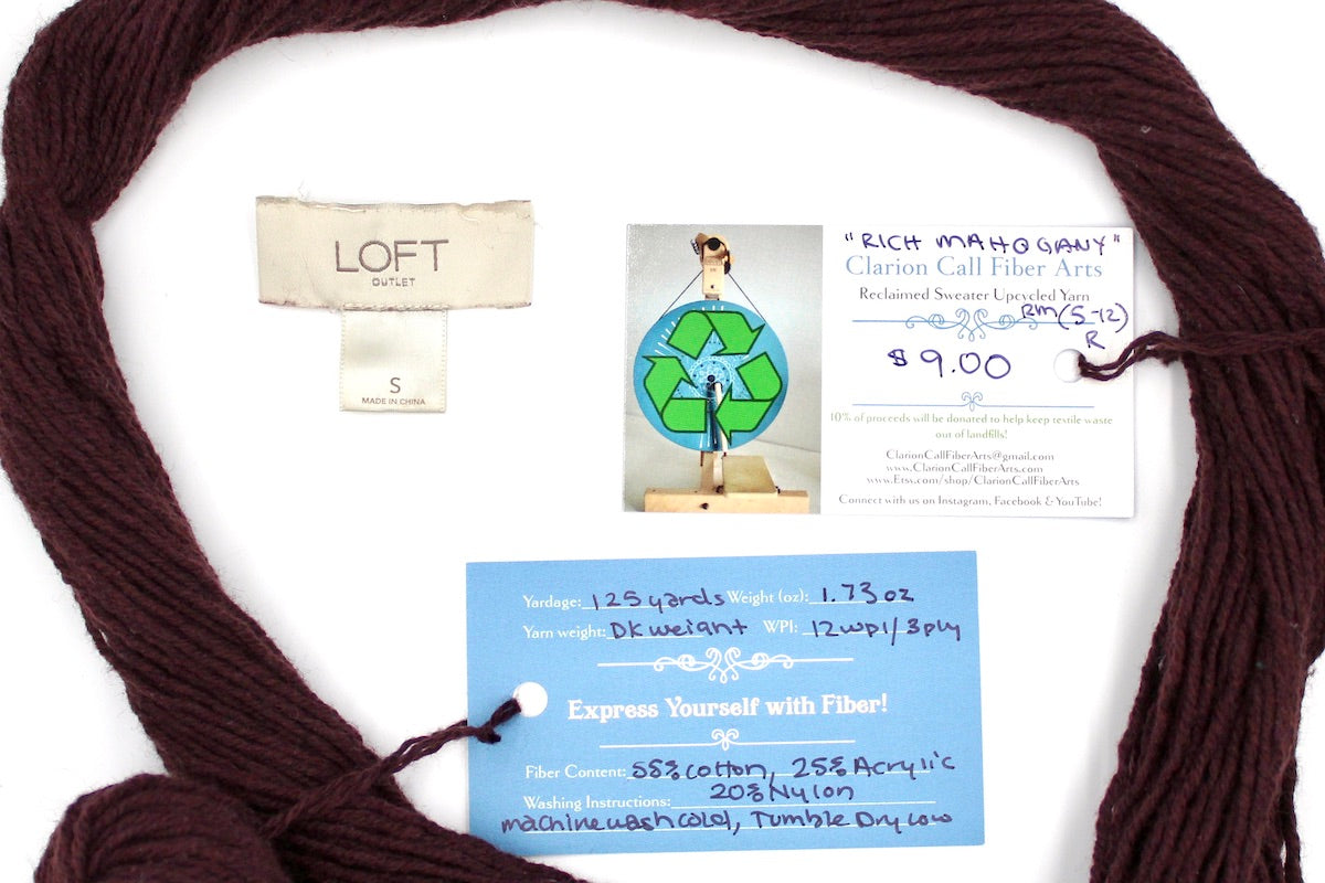 A skein of Vegan, Maroon/ Reddish Brown, Cotton/ Acrylic/ Nylon, Dk Weight yarn encircling the sweater labels it was recycled from, as well as the hand written yarn tags detailing the specs of this upcycled yarn to help you make your next project!