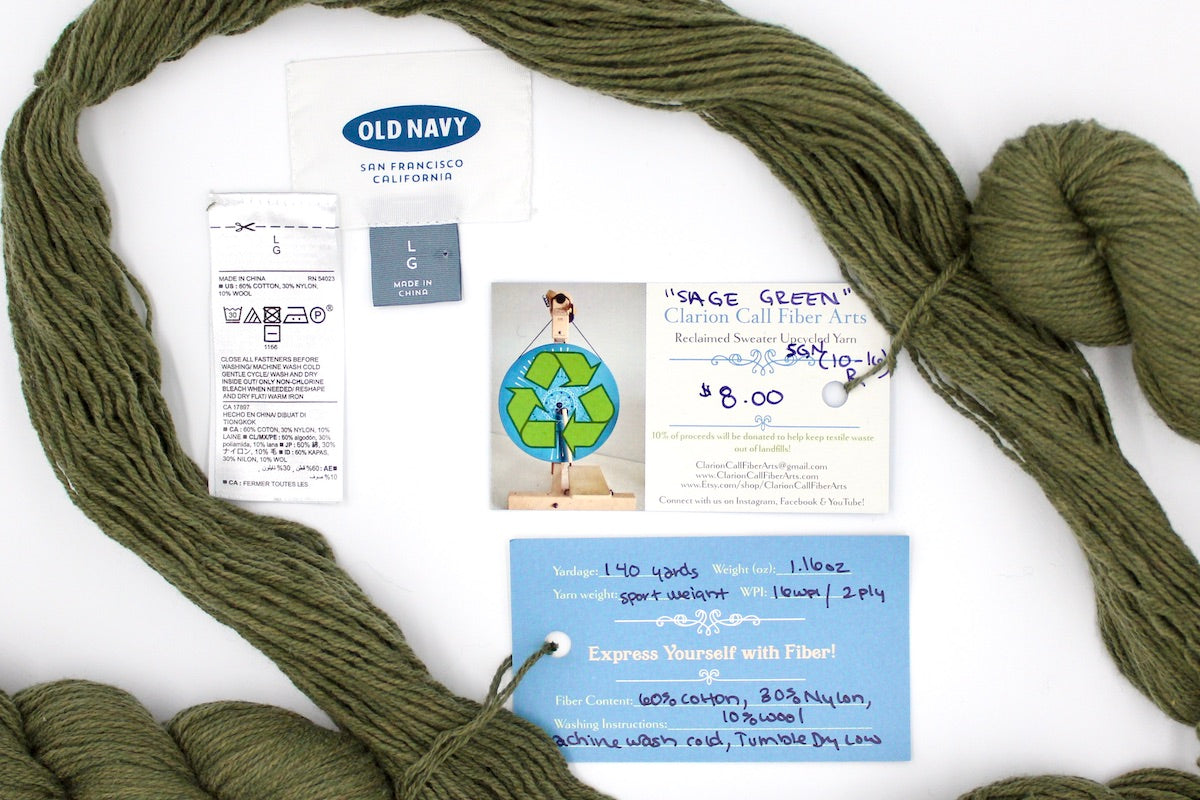 A skein of Heathered Sage, Olive Green, Cotton/ Nylon/ Wool, Sport Weight yarn encircling the sweater labels it was recycled from, as well as the hand written yarn tags detailing the specs of this upcycled yarn to help you make your next project!