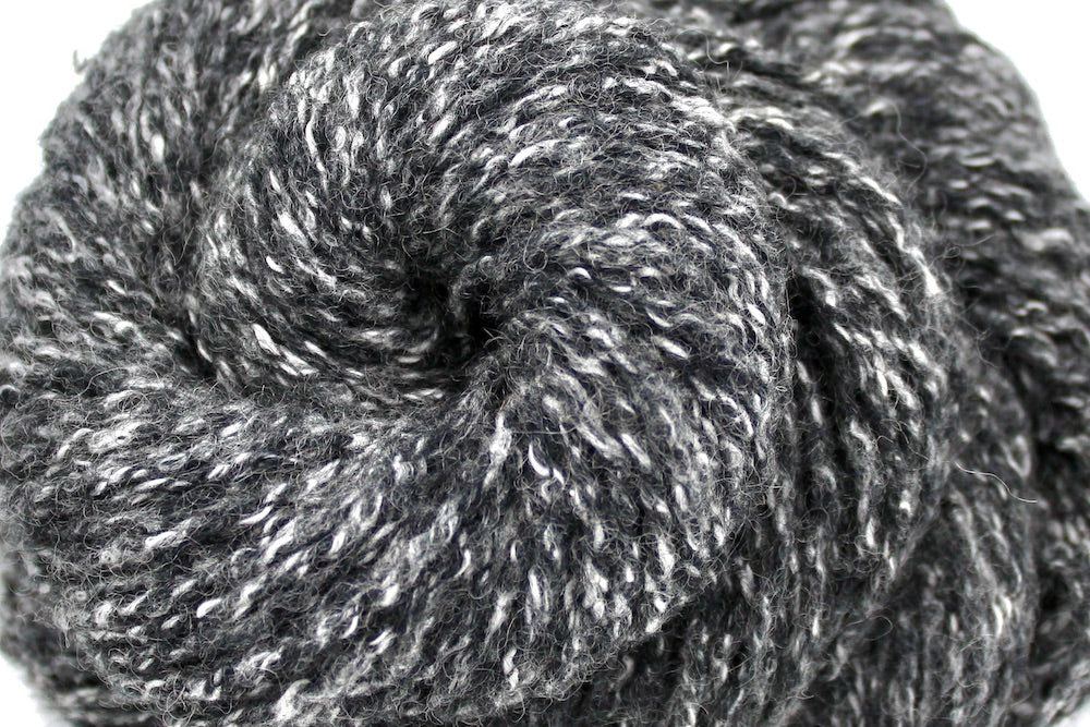 A close up shot of a skein of Heathered Charcoal Grey and White, Wool/ Acrylic/ Cotton/ Polyester, Heavy Worsted weight Yarn recycled by hand from unwanted sweaters beautifully coiled in the center of the frame.