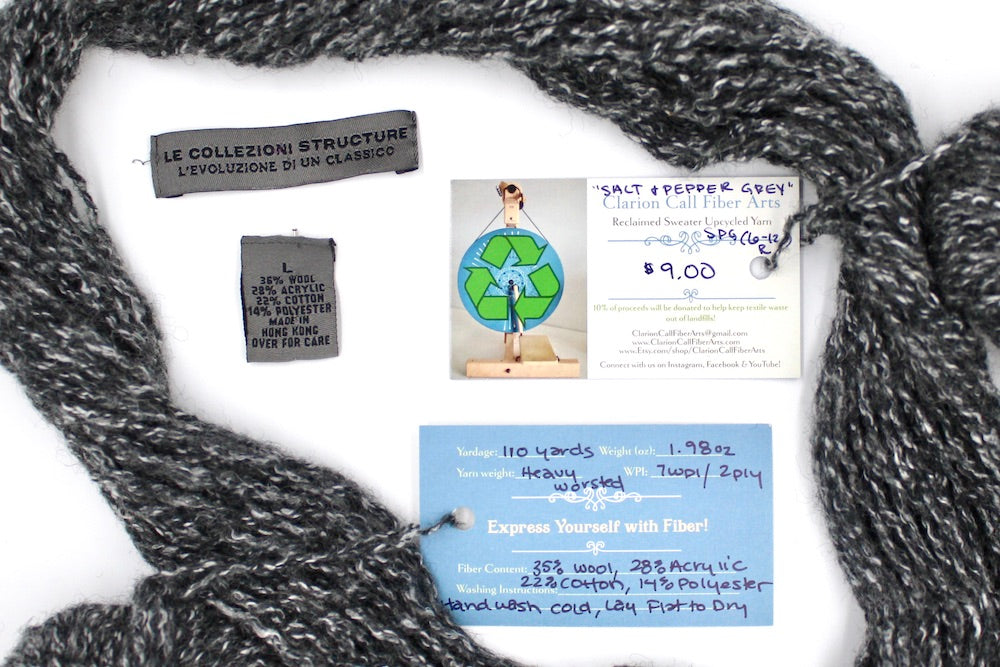 A skein of Heathered Charcoal Grey and White, Wool/ Acrylic/ Cotton/ Polyester, Heavy Worsted Weight yarn encircling the sweater labels it was recycled from, as well as the hand written yarn tags detailing the specs of this upcycled yarn to help you make your next project!