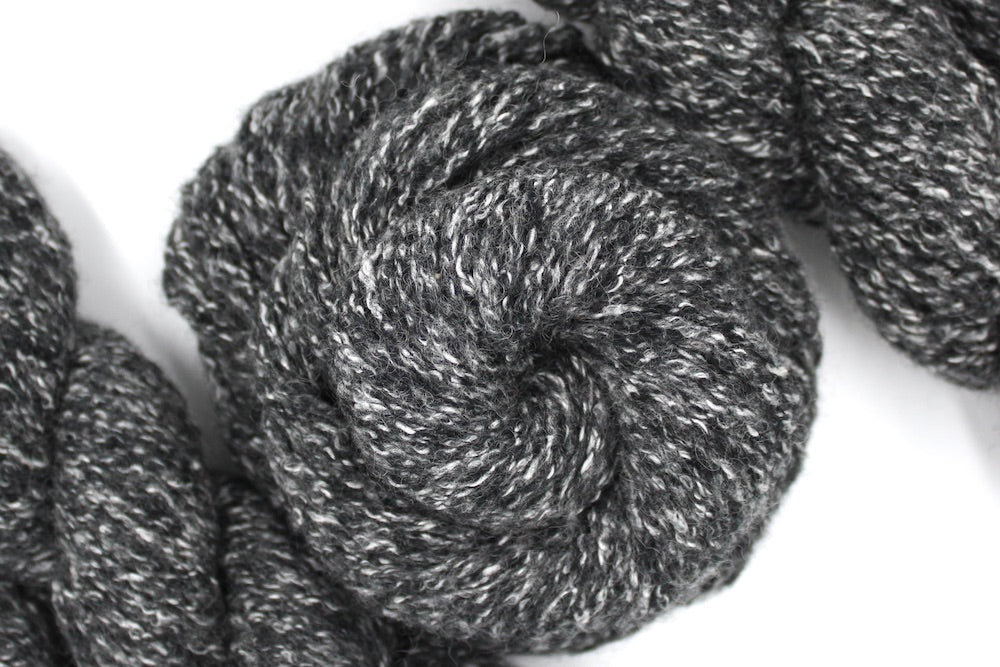 A skein of Heathered Charcoal Grey and White, Wool/ Acrylic/ Cotton/ Polyester, Heavy Worsted weight Yarn recycled by hand from unwanted sweaters swirled attractively in the center of the frame.