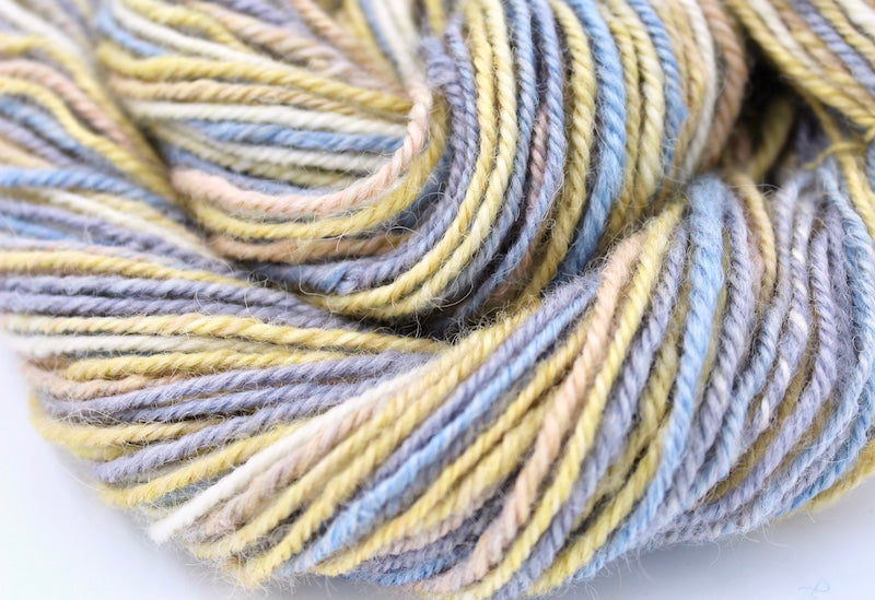 A close up view of a one of a kind, hand dyed variegated skein of multicolored Grey, Baby Blue, Pastel Pink, Yellow, and White self-striping wool Yarn.