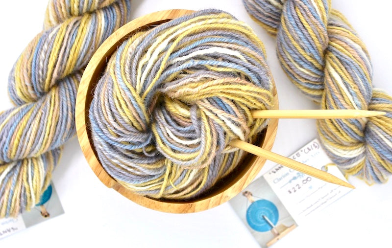 A one of a kind, hand dyed variegated skein of multicolored Grey, Baby Blue, Pastel Pink, Yellow, and White self-striping wool Yarn, in a yarn bowl with knitting needles, ready to be made into something beautiful!