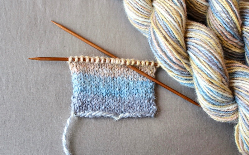 A sample swatch knitted from a one of a kind, hand dyed multicolored Grey, Baby Blue, Pastel Pink, Yellow, and White self-striping wool Yarn.