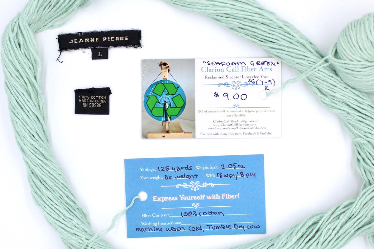 A skein of Vegan, Seafoam/ Aquamarine Blueish Green, 100% Cotton, Dk Weight yarn encircling the sweater labels it was recycled from, as well as the hand written yarn tags detailing the specs of this upcycled yarn to help you make your next project!
