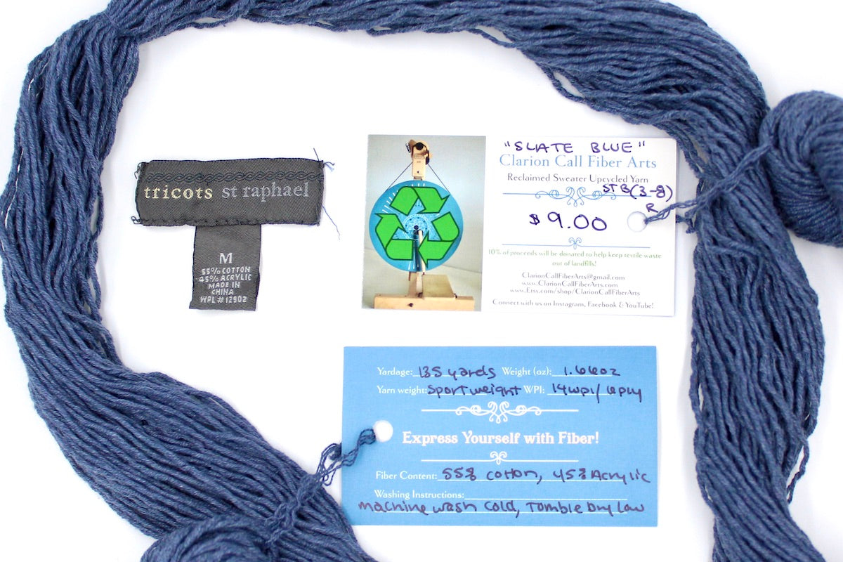 A skein of Vegan, Heathered Slate Grayish Blue, Cotton/ Acrylic, Sport Weight yarn encircling the sweater labels it was recycled from, as well as the hand written yarn tags detailing the specs of this upcycled yarn to help you make your next project!