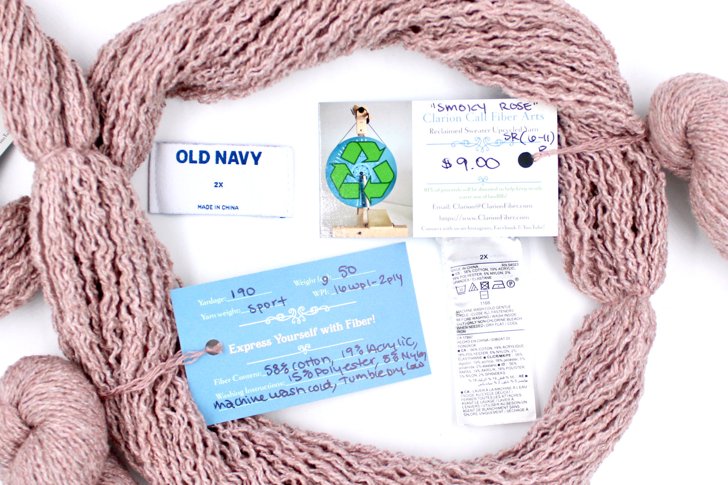 A skein of Vegan, Dusky Pink, Cotton, Polyester, Acrylic, Nylon, Sport Weight yarn encircling the sweater labels it was recycled from, as well as the hand written yarn tags detailing the specs of this recycled yarn to help you make your next project!