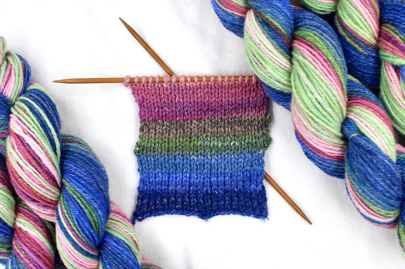 A sample swatch knitted from a one of a kind, hand dyed gradient skein of multicolored Navy Blue, Royal Blue, Teal, Green, Olive Green, Magenta, Salmon, Pastel Pink and Mint Green self-striping wool Yarn.
