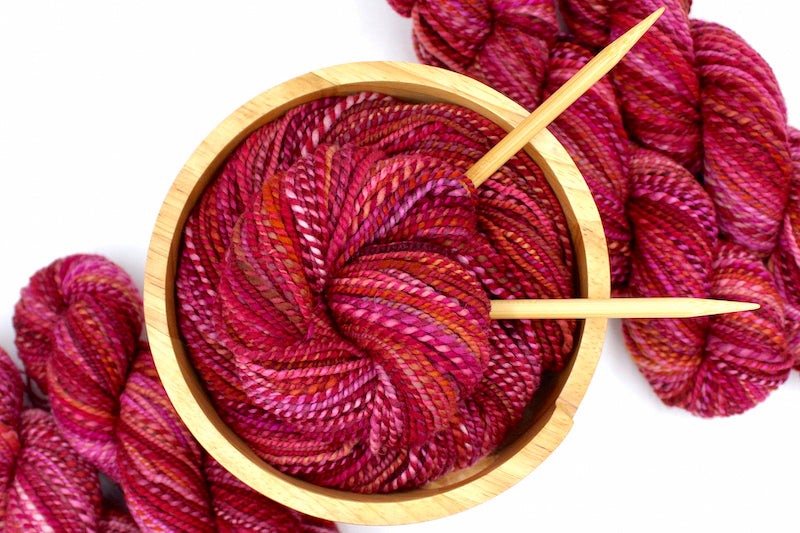 A one of a kind, hand dyed variegated skein of multicolored Red, Pink, and Orange self-striping wool Yarn, in a yarn bowl with knitting needles, ready to be made into something beautiful!