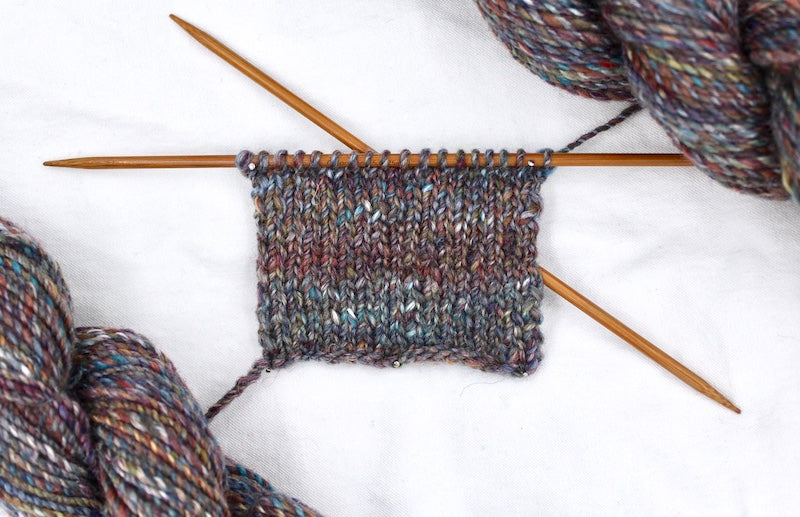 A sample swatch knitted from a one of a kind, hand dyed variegated skein of multicolored Maroon, Red, Grey, Blue, Yellow, and Brown self-striping wool Yarn.