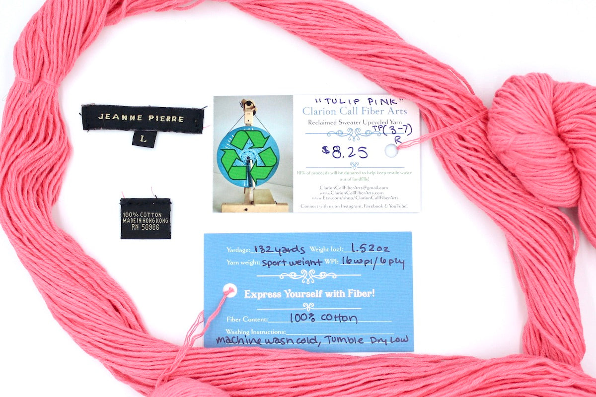 A skein of Vegan, Neon Rose Pink/ Fuchsia, 100% Cotton, Sport Weight yarn encircling the sweater labels it was recycled from, as well as the hand written yarn tags detailing the specs of this upcycled yarn to help you make your next project!