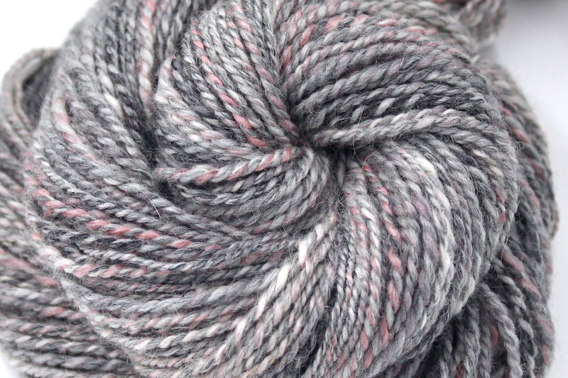 A close up view of a one of a kind, hand dyed variegated skein of multicolored Grey, White, and Pink self-striping wool Yarn.