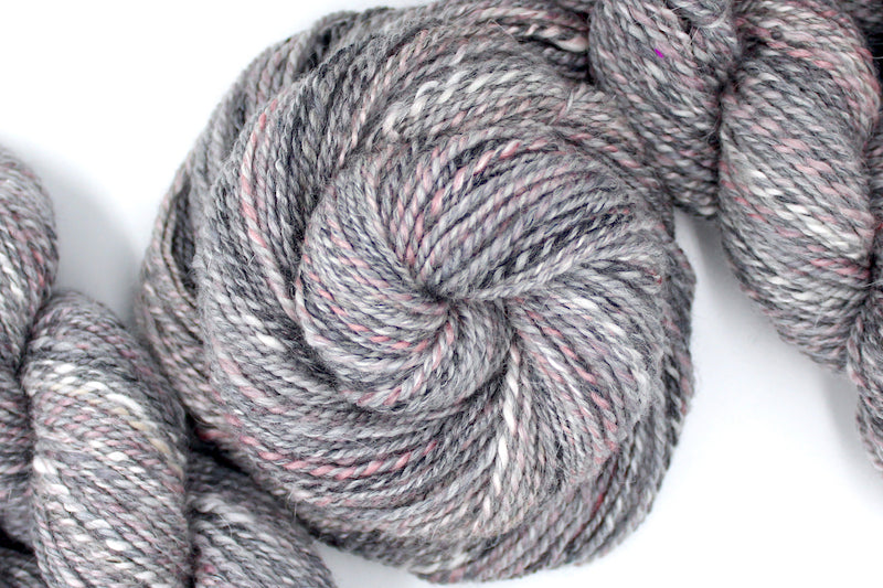 A one of a kind, hand dyed variegated skein of multicolored Grey, White, and Pink self-striping wool Yarn coiled attractively in the center of the frame.
