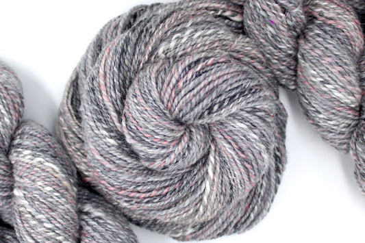 A one of a kind, hand dyed variegated skein of multicolored Grey, White, and Pink self-striping wool Yarn coiled attractively in the center of the frame.