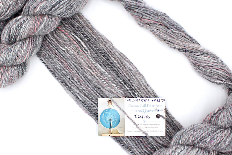 A one of a kind, hand dyed variegated skein of multicolored Grey, White, and Pink self-striping wool Yarn draped diagonally across the frame, so you can really see the color play.