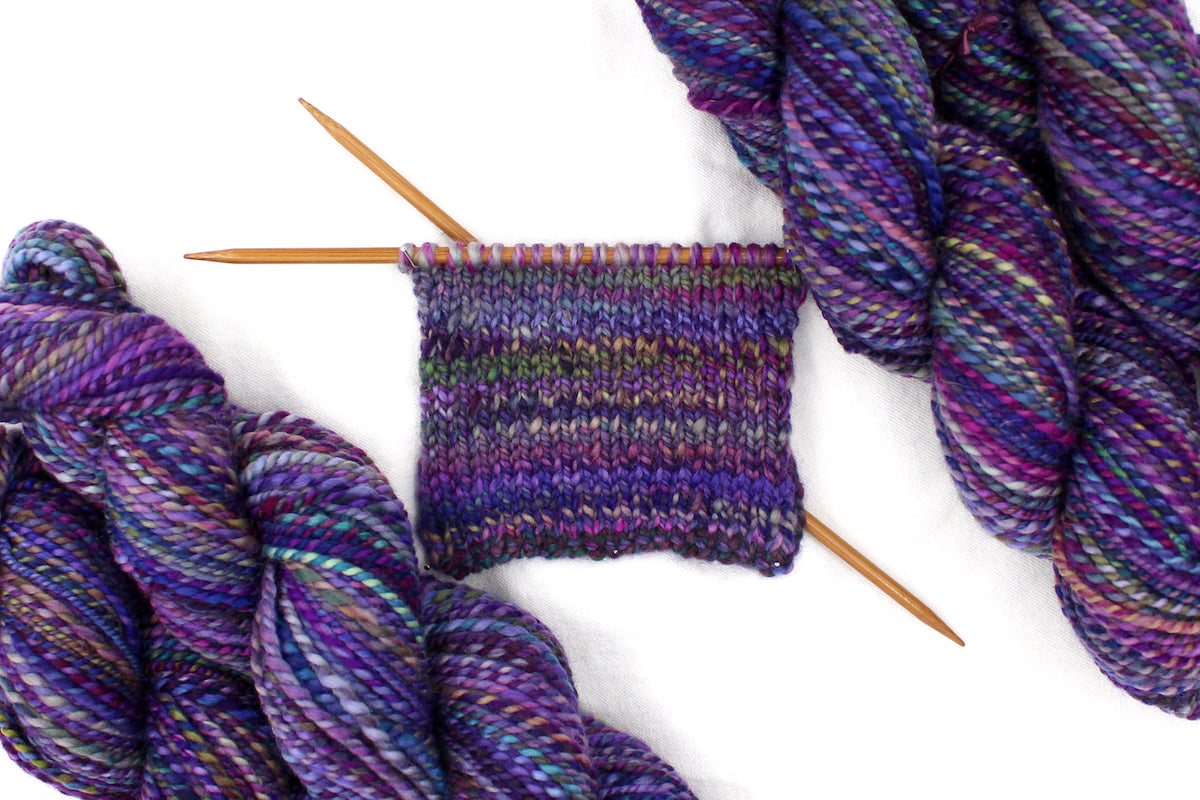 A sample swatch knitted from a one of a kind, Worsted weight, hand dyed variegated skein of multicolored Purple, Fuchsia, Green, Navy Blue, Teal, and Pink self-striping wool Yarn.