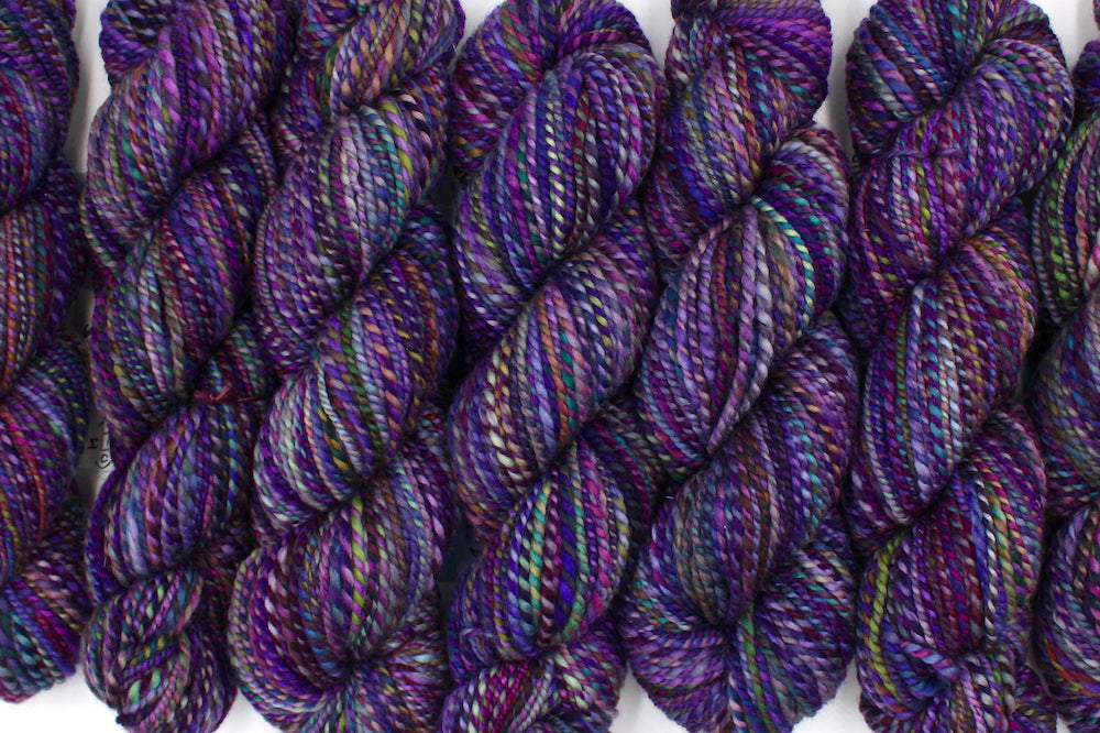 Six one of a kind, Worsted weight, hand dyed variegated skein of multicolored Purple, Fuchsia, Green, Navy Blue, Teal, and Pink self-striping wool Yarn lined up side by side.