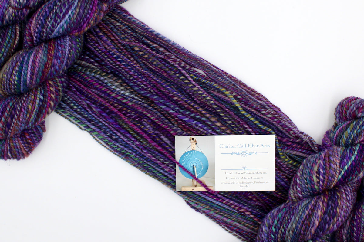A one of a kind, Worsted weight, hand dyed variegated skein of multicolored Purple, Fuchsia, Green, Navy Blue, Teal, and Pink self-striping wool Yarn draped diagonally across the frame, so you can really see the color play.