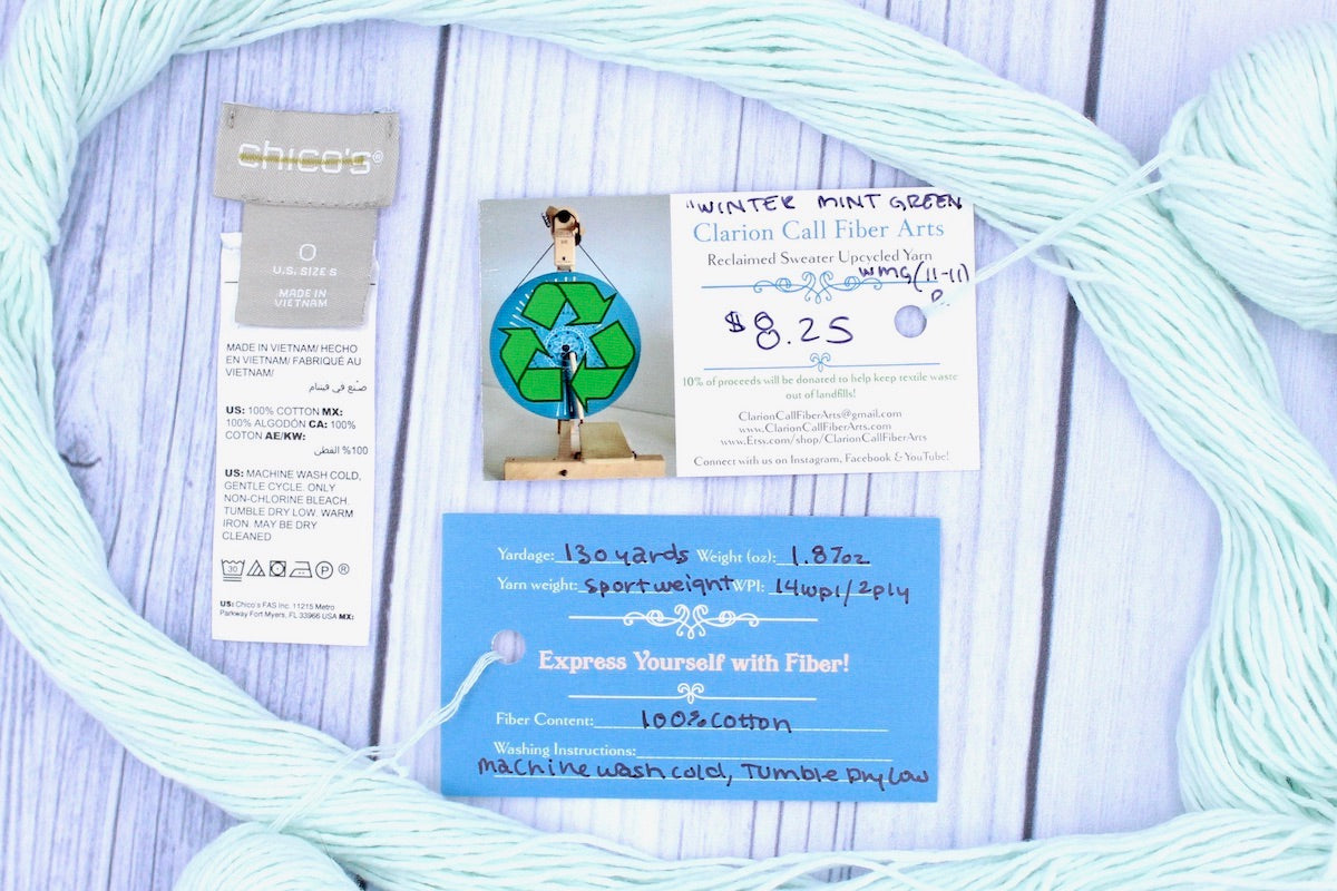 A skein of Vegan, Light Pastel Mint Green, 100% Cotton, Sport Weight yarn encircling the sweater labels it was recycled from, as well as the hand written yarn tags detailing the specs of this upcycled yarn to help you make your next project!