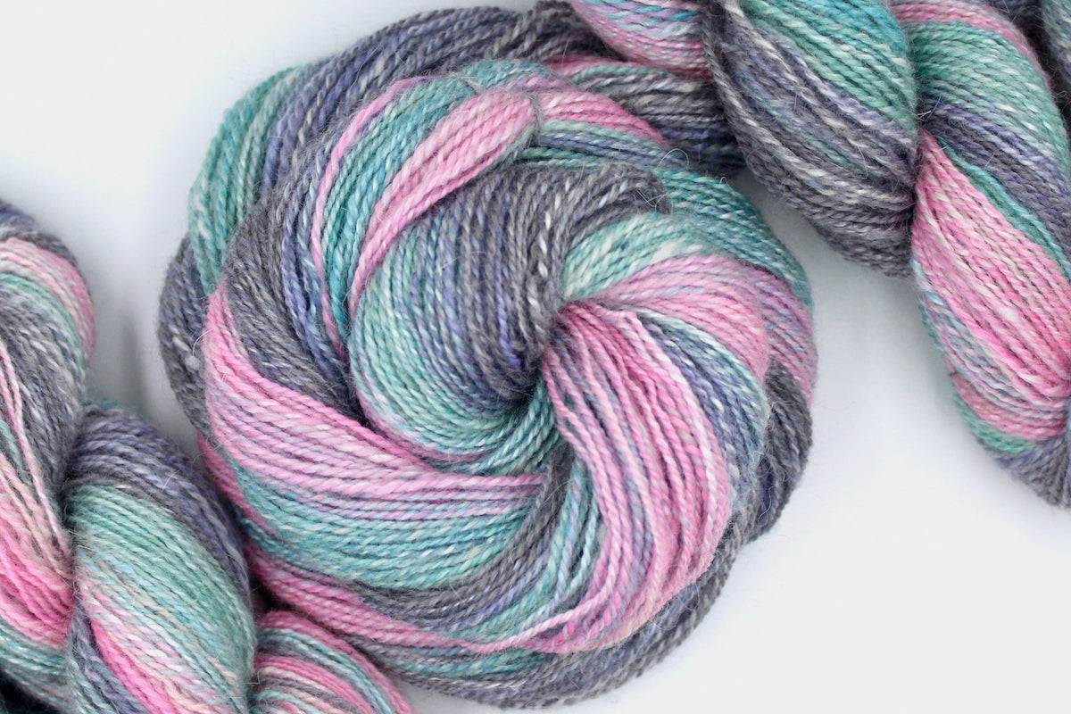 A one of a kind, hand dyed gradient skein of multicolored Light Pink, Sky Blue, Seafoam Green, Grey, and Lavender self-striping wool Yarn coiled attractively in the center of the frame. 