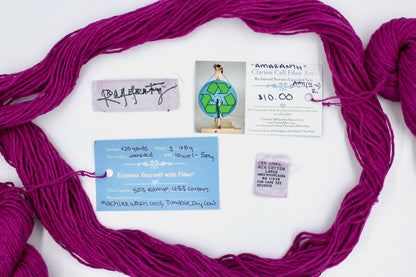 A skein of Vegan, Bright Fuchsia Pink, 55% Ramie, 45% Cotton, Worsted Weight yarn encircling the sweater labels it was recycled from, as well as the hand written yarn tags detailing the specs of this recycled yarn to help you make your next project! 