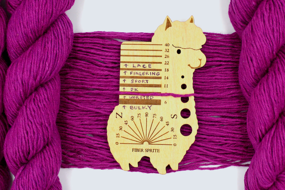 A skein of Vegan, Bright Fuchsia Pink, 55% Ramie, 45% Cotton, Worsted Weight Yarn recycled by hand from unwanted sweaters draped over the center of the frame with one strand on top of a Llama shaped Yarn weight tool, to better illustrate the thickness of the yarn. 