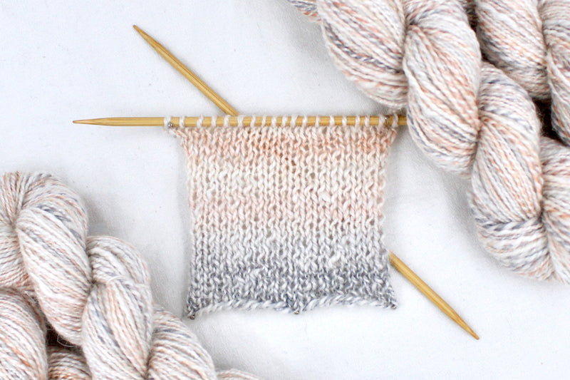 A sample swatch knitted from a one of a kind, hand dyed variegated skein of multicolored Cream, Dusky Pink, and Grey self-striping wool Yarn. 